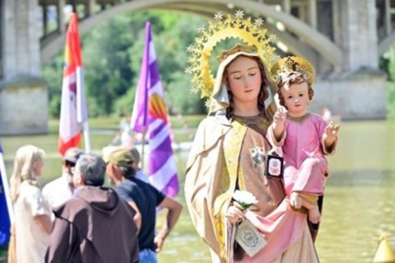 Religious statue of Mary and child near a river with people and flags.